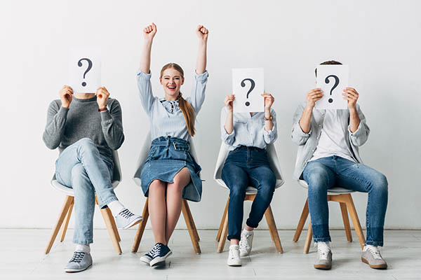 Image shows 4 job seekers sitting in chairs. Three of them are holding question mark signs over their faces, and one woman is raising her hands in celebration.