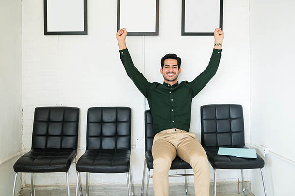 Image shows a male job seeker sitting in a chair in a waiting room, celebrating with his hands up.
