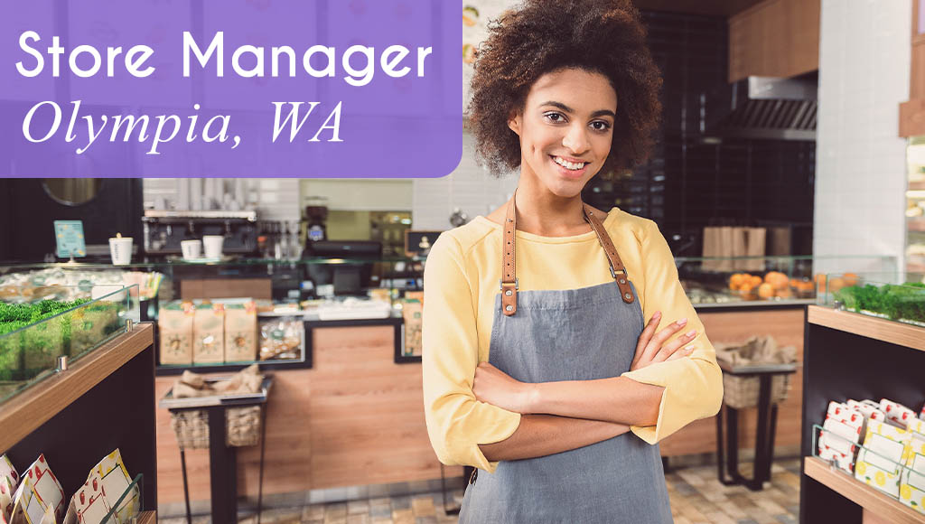 Image shows a woman smiling and wearing an apron standing in a store front next to shelving lined with products. Behind her is a store counter with a cash register and more store products. Now Hiring a Store Manager in Olympia, WA. All StarZ Staffing.