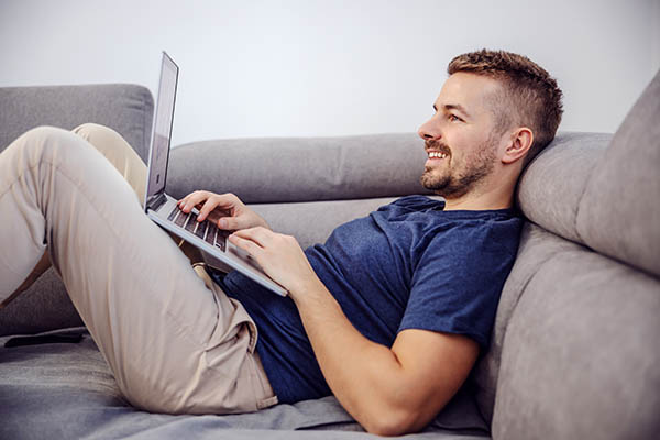 Image shows a smiling man lying on gray sofa, using a laptop to research before an interview.