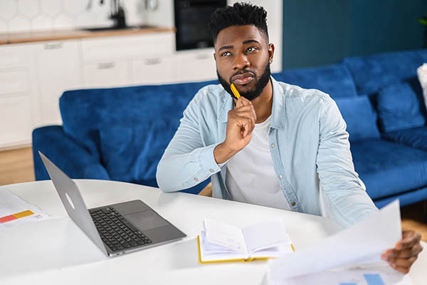 Image shows a young man holding his resume with a thoughtful look on his face. He is sitting at a table with a laptop. There is a blue couch in the background. All StarZ Staffing.