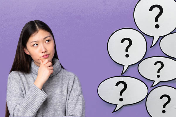 A woman is touching her face with an inquisitive look on her face in front of a purple background with speech bubbles containing question marks.