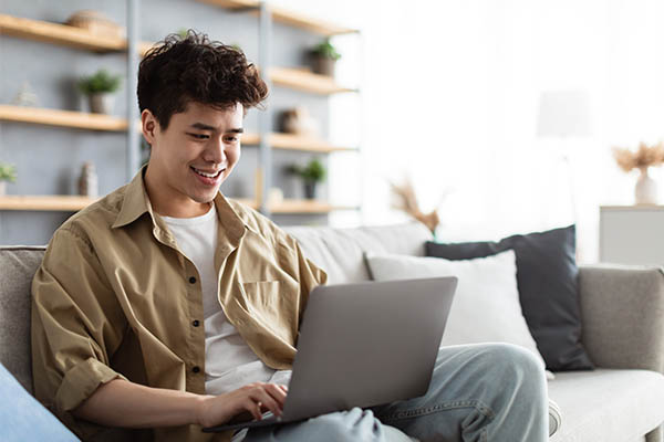 A male job seeker is smiling and typing on a laptop, researching the company ahead of a job interview.