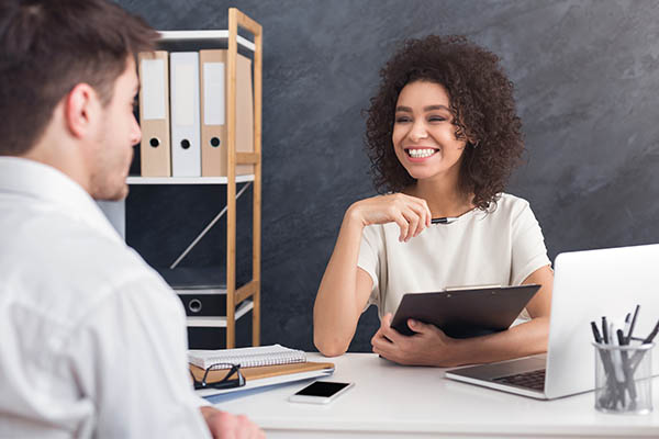 Image shows a man speaking to a smiling female recruiter. She is holding a clipboard. They are sitting at a desk in an office setting.