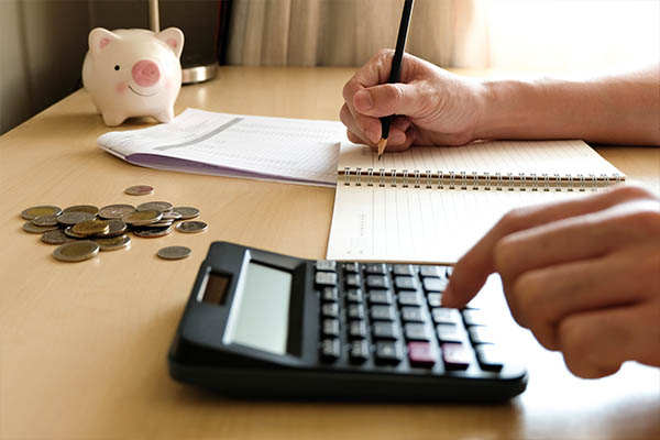Image shows hands writing on a notepad and using a calculator to anaylze the cost savings of a second-chance employment program. A piggy bank and a pile of coins are on the desk next to the notepad.
